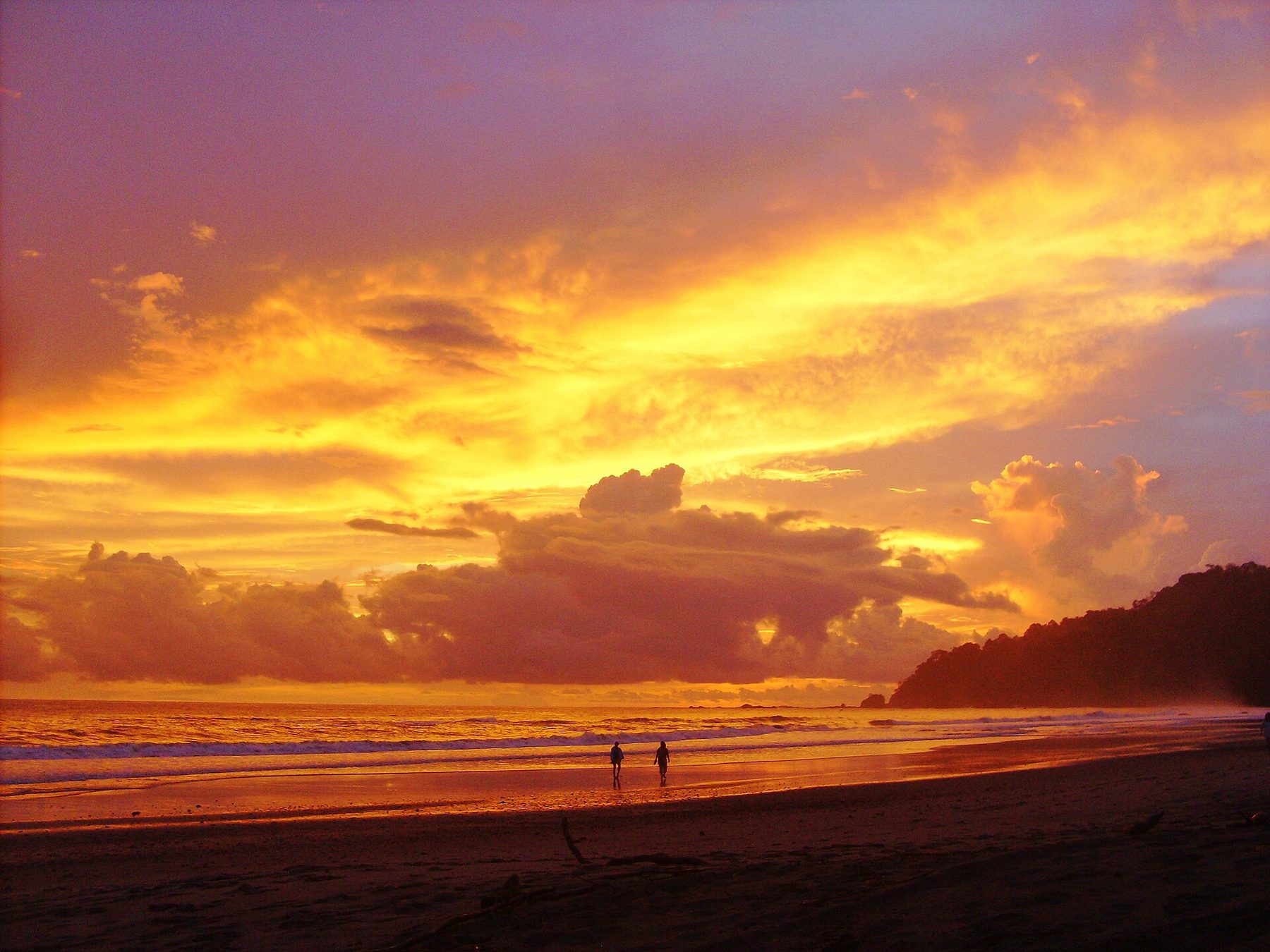 Two people walk on wet sand at Playa Espadilla under an orange and gold sunset, with a forested headland on the right