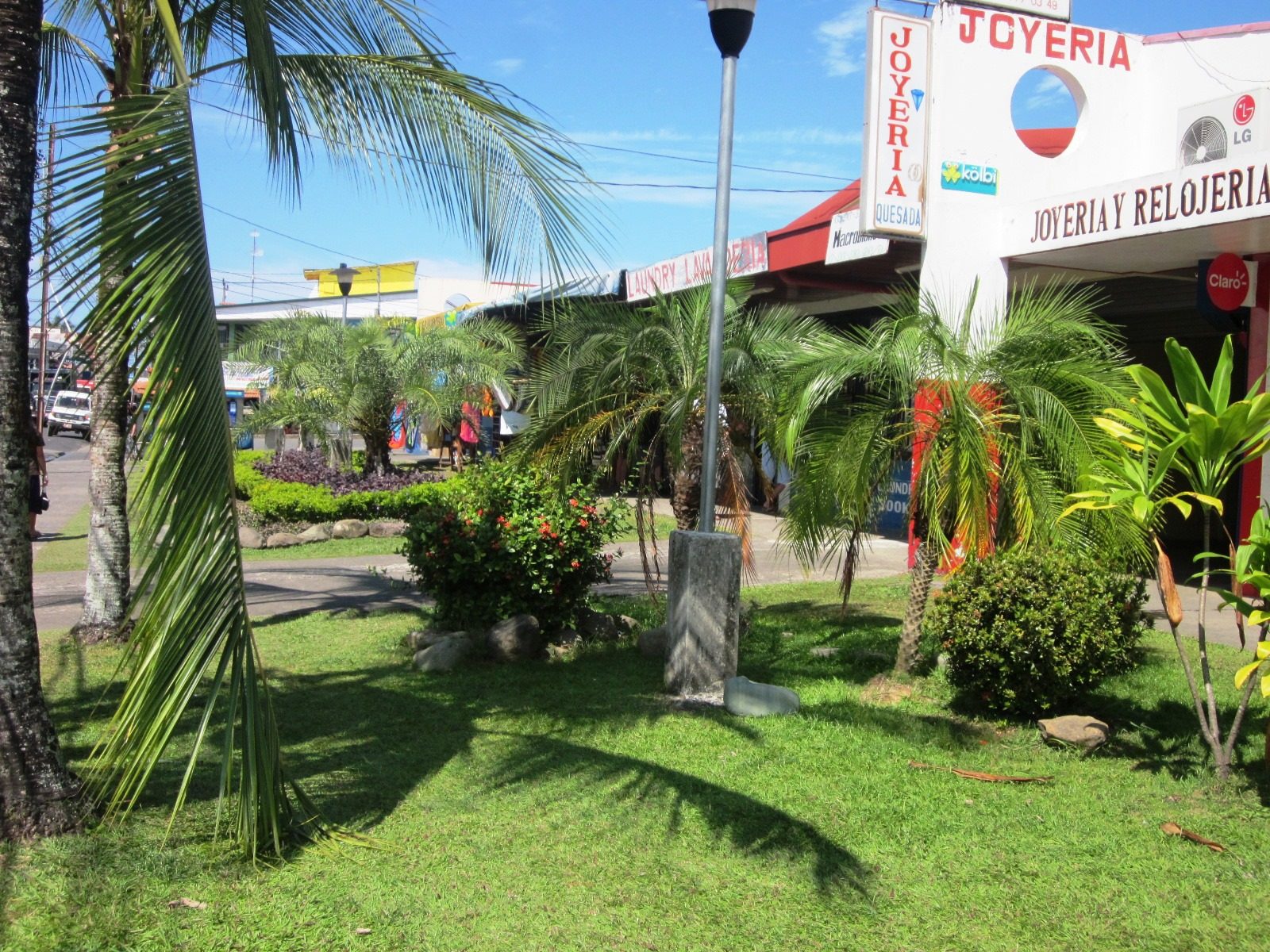 Palms, grass and low shopfronts beside the municipal market in Quepos, with a jewellery shop and a laundry sign on the corner