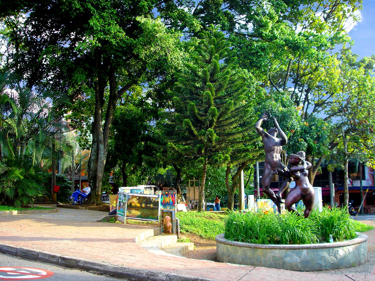 A tree-shaded plaza ringed by low buildings and bar frontages at Parque Lleras in El Poblado, Medellín