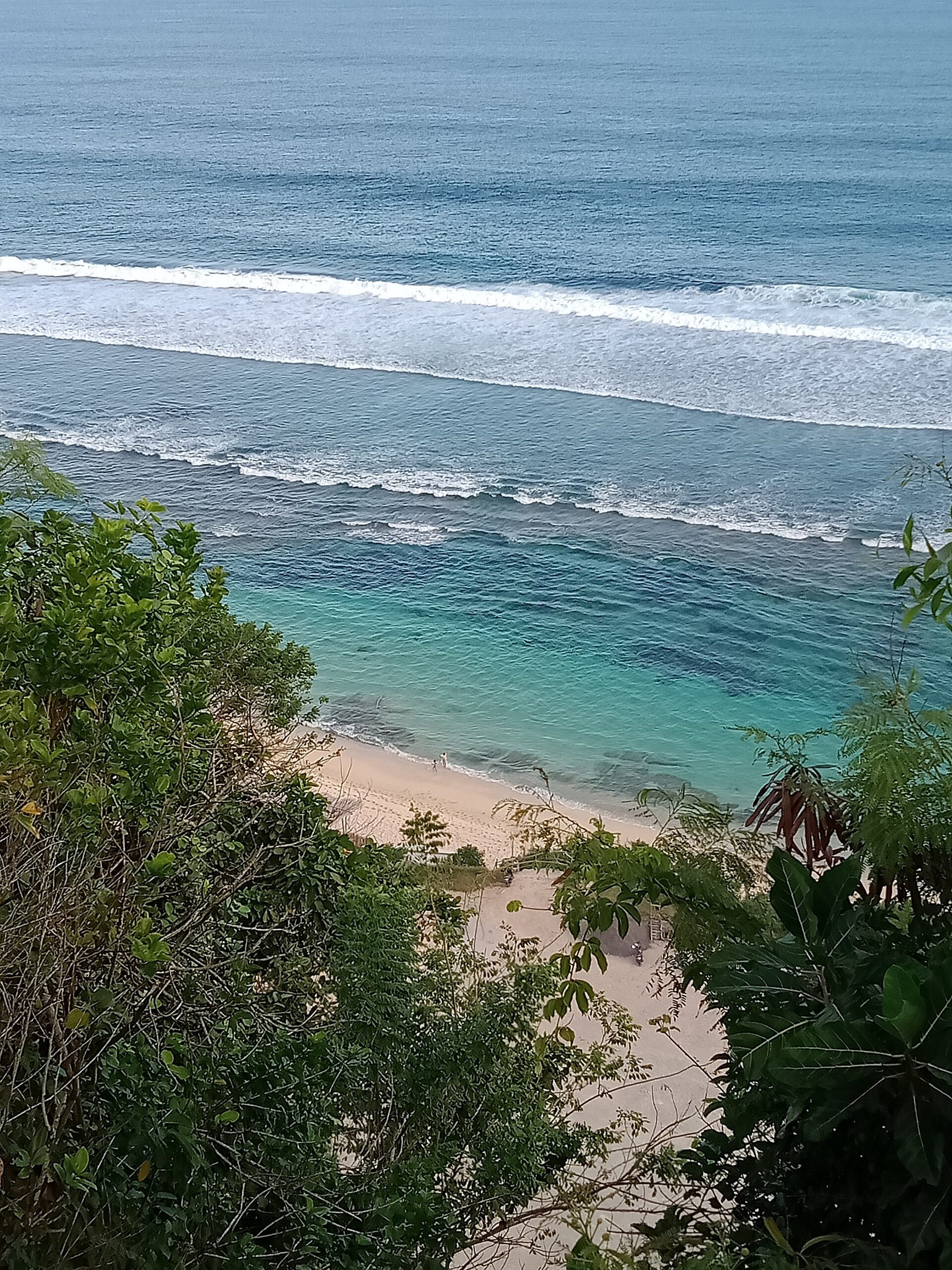 A view down from the clifftop through green foliage onto a straight strip of pale sand, two people walking at the waterline, and lines of surf beyond