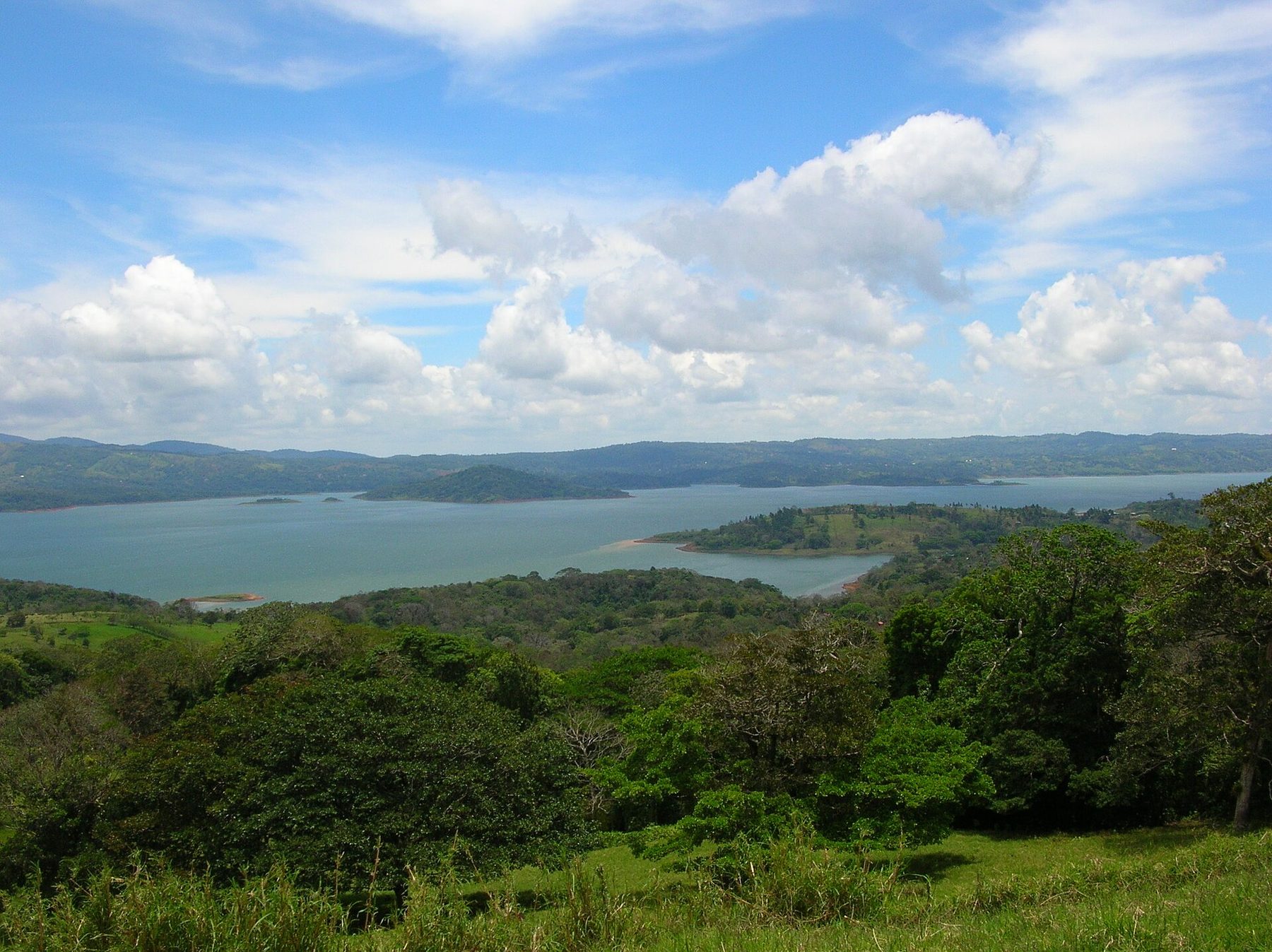 A wide grey-blue lake fills a green valley below rolling pasture and forested ridges, with low cloud sitting on the hills along the far shore