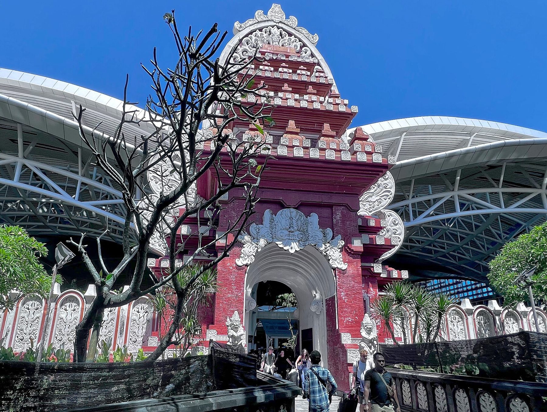 Travelers with bags walk out through the tall red brick and white carved Balinese gate at the entrance to Ngurah Rai airport, with the terminal's curved steel canopy behind it under a blue sky