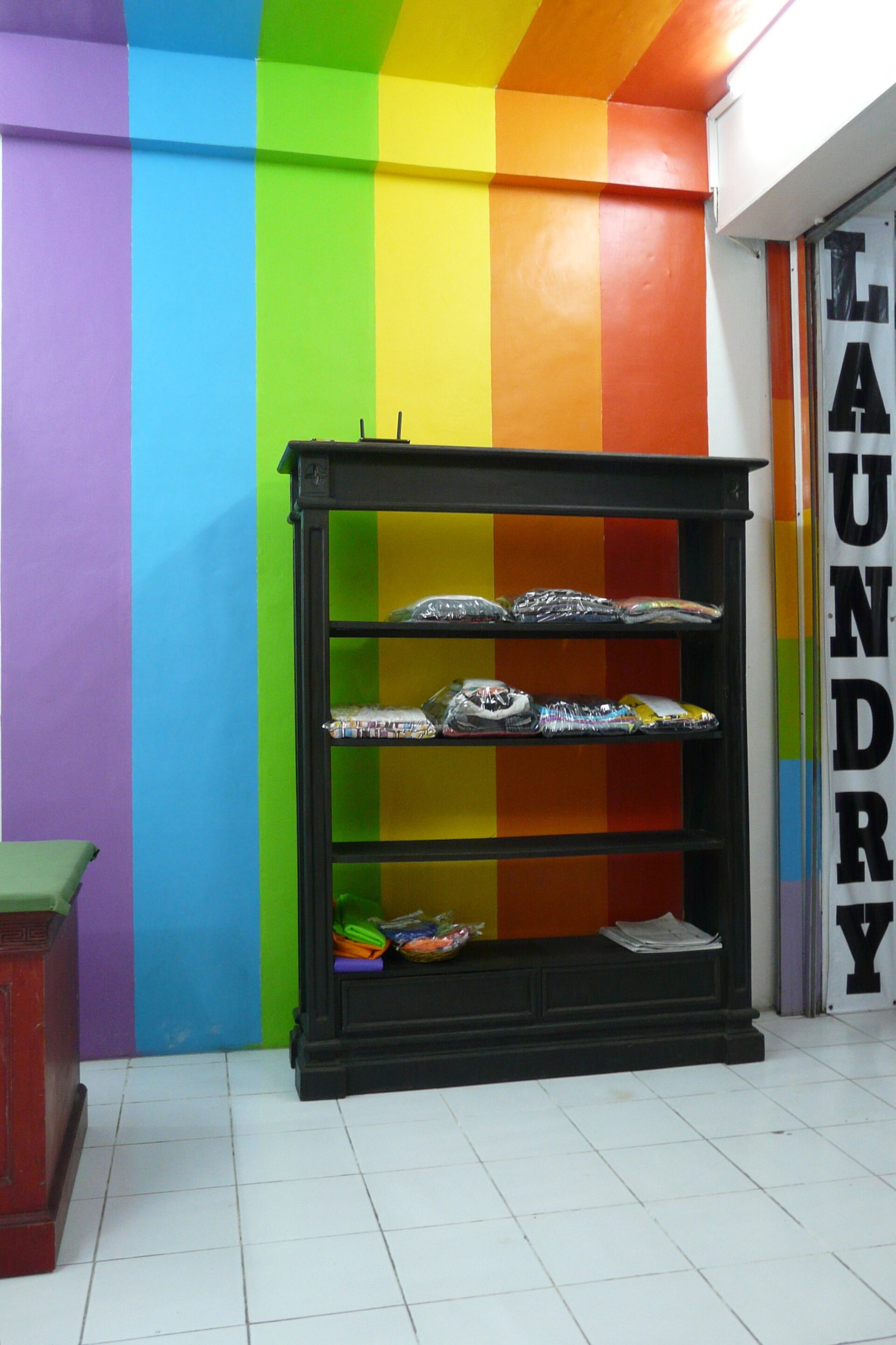 The inside of a small laundry shop with a rainbow-striped wall, a black shelf unit holding folded clothes bagged in clear plastic, and a vertical Laundry sign by the door
