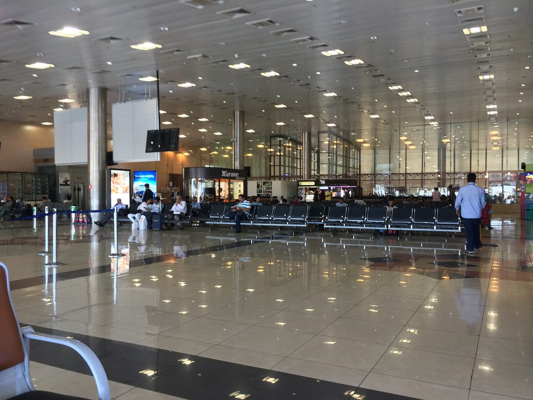 A wide airport departure hall with long rows of steel and blue seating, a polished tiled floor and a handful of waiting passengers