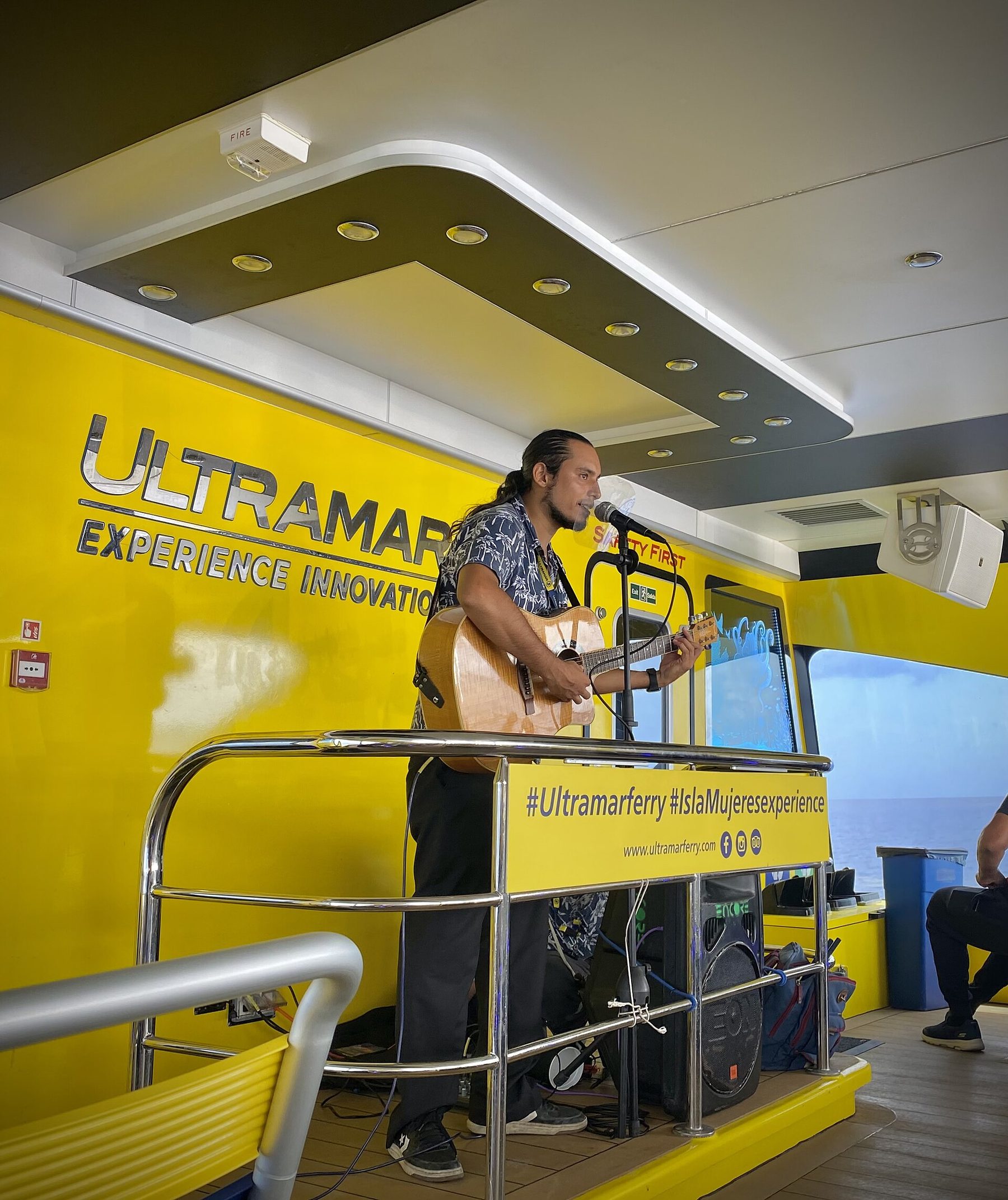 Passengers seated on the open upper deck of a catamaran ferry crossing between Cozumel and Playa del Carmen, with a guitarist playing.