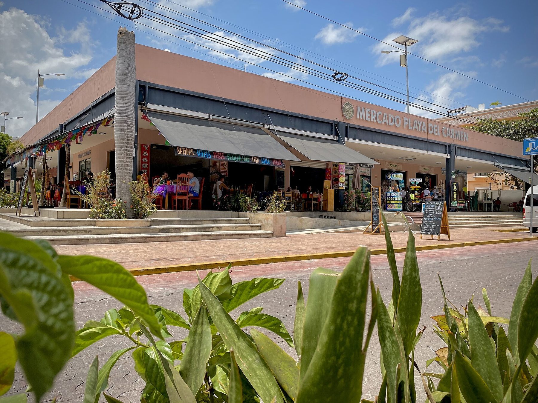 A low pink market building signed Mercado Playa del Carmen, with open-front restaurants under awnings, chalkboard menus on the sidewalk and diners at wooden tables