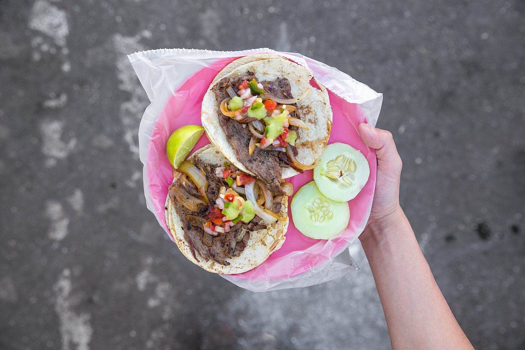 A hand holds a pink plastic plate lined with a bag, carrying two beef tacos topped with onions, pico de gallo and green salsa, beside a lime wedge and cucumber slices