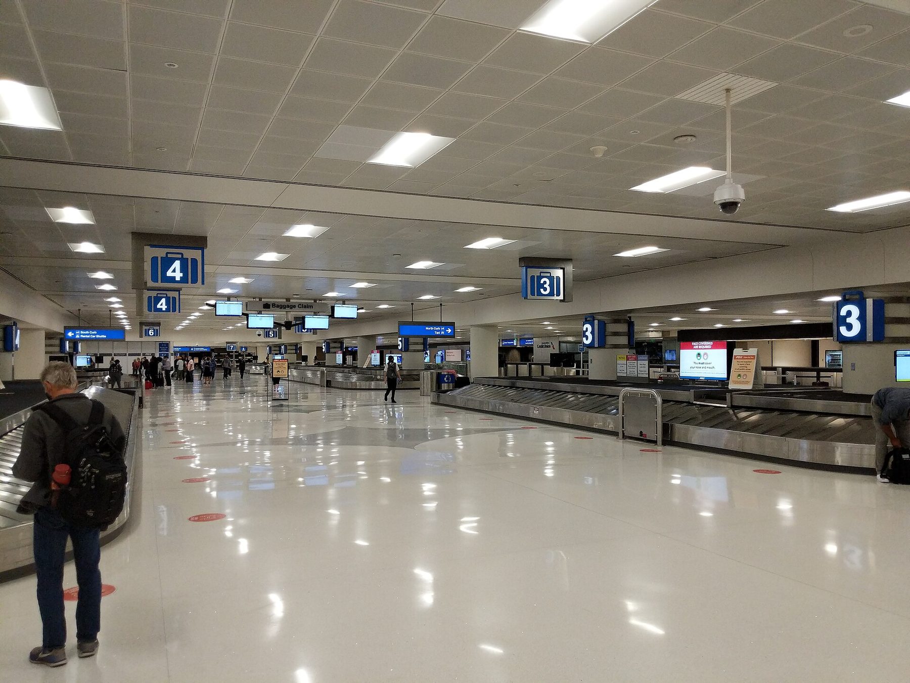 Travelers wait beside a curved metal baggage carousel in a tall airport hall with a patterned tile floor