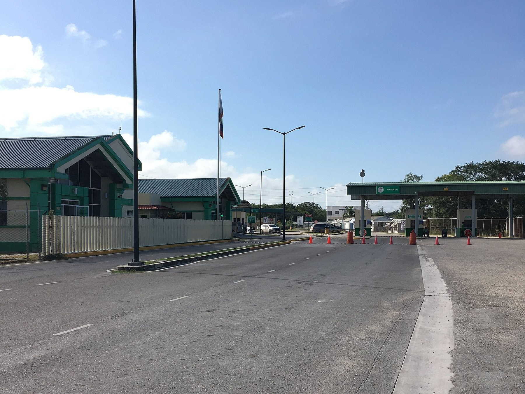 A quiet border post with green-roofed immigration buildings, a Belize flag on a tall pole and orange traffic cones across an empty road