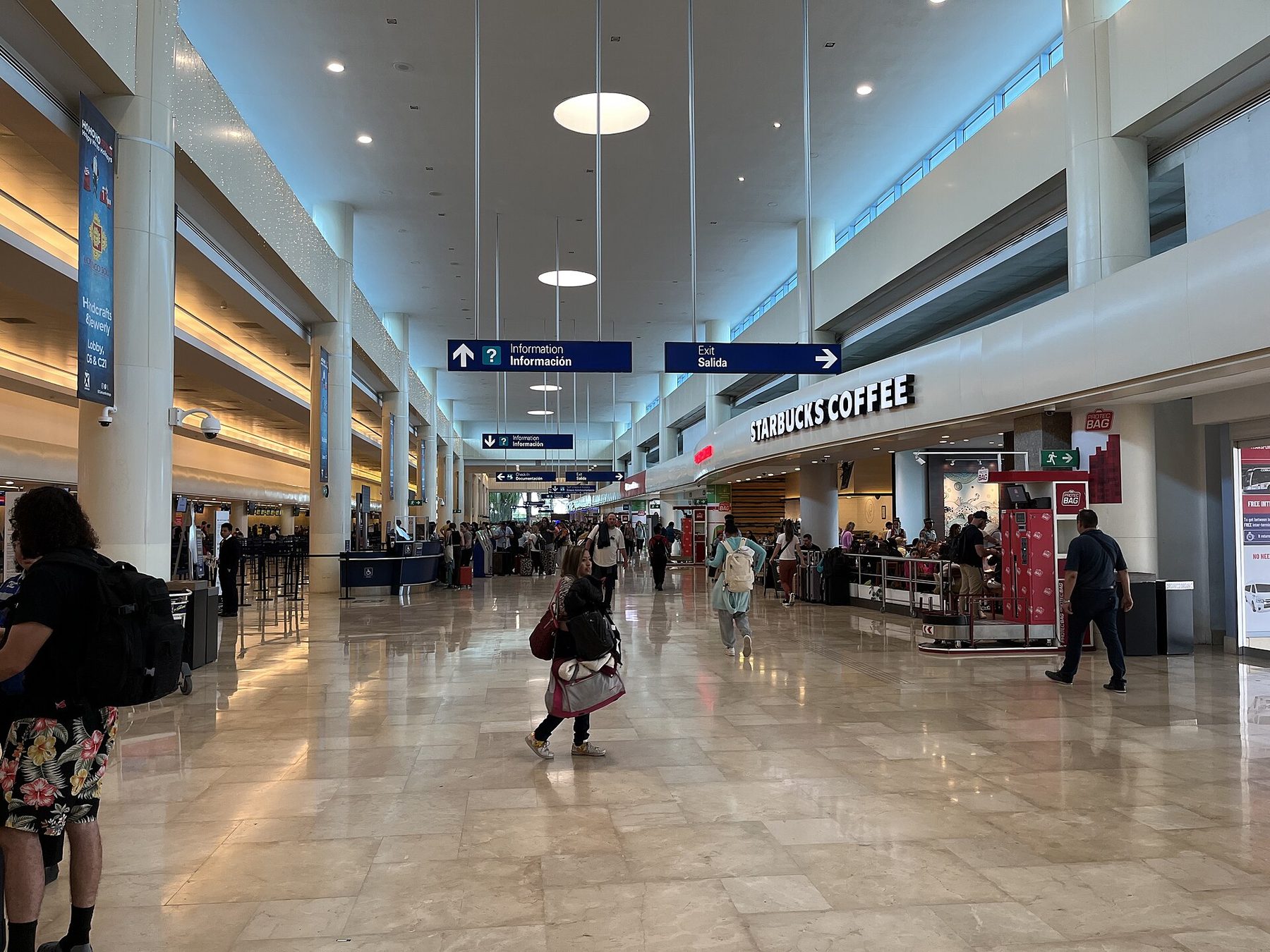 Travelers with backpacks and rolling bags cross the wide marble concourse of a bright airport terminal, under blue signs reading Information and Exit