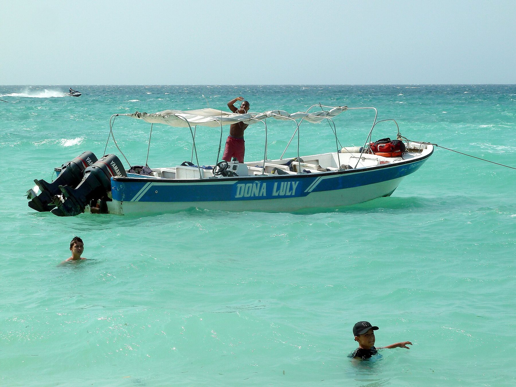 A wooden passenger boat with an outboard motor and a blue canopy floats in shallow clear water off a sandy beach