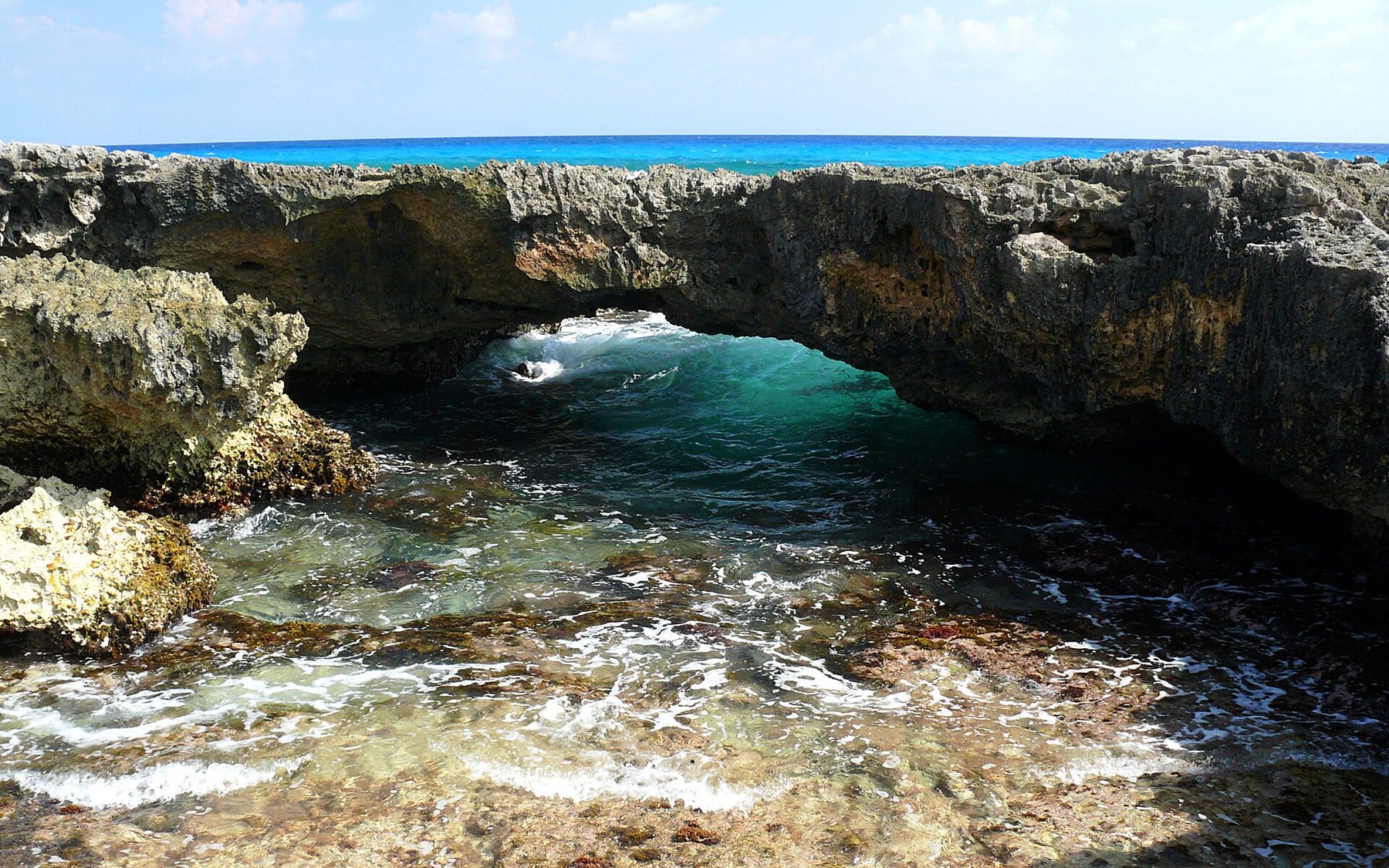 Waves break over a broken, shallow reef along an exposed rocky shoreline under a bright sky