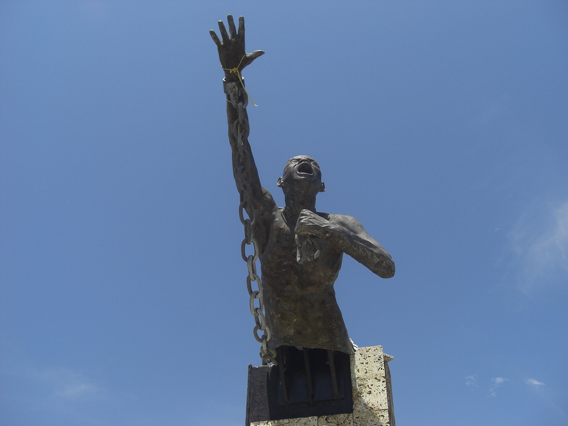 A bronze statue of a man with one arm raised and a broken chain hanging from his wrist, head back and mouth open, standing on a stone plinth against a clear blue sky