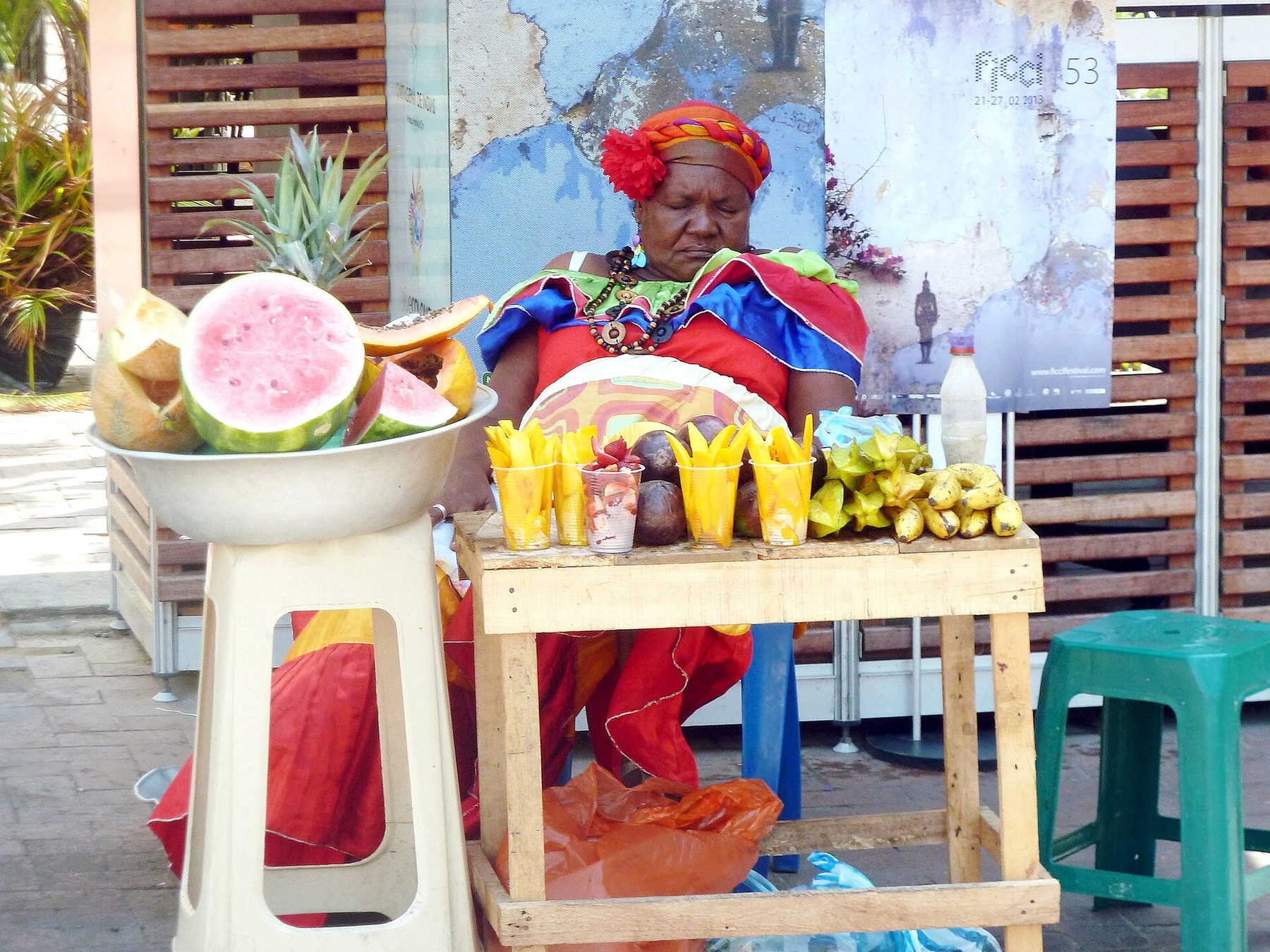A woman in a red, blue and green ruffled dress and a striped headwrap sits behind a wooden stand of sliced watermelon, papaya, mango cups, bananas and star fruit