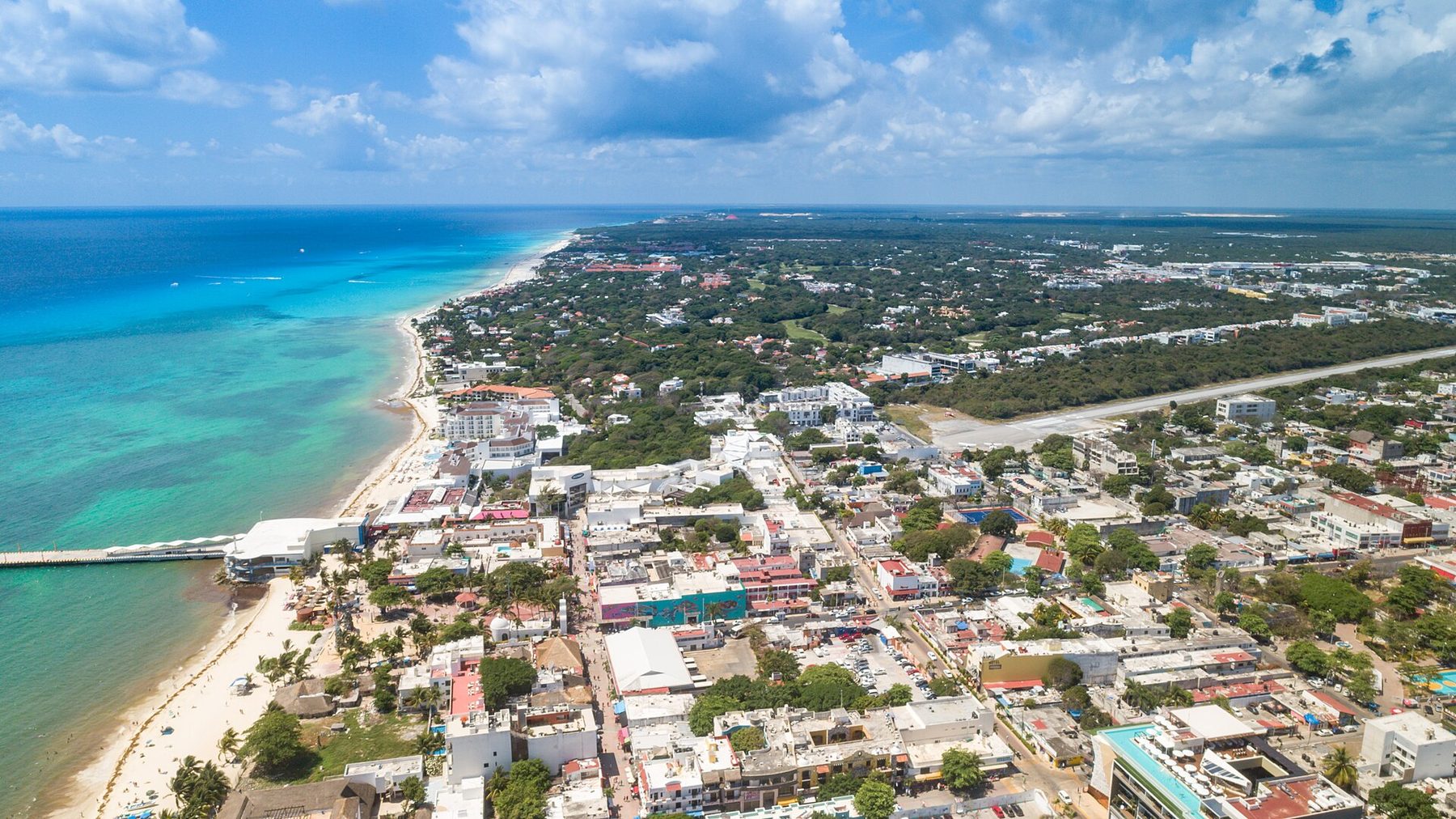 An aerial view of the Playa del Carmen coast, turquoise water meeting white sand and low buildings among palms