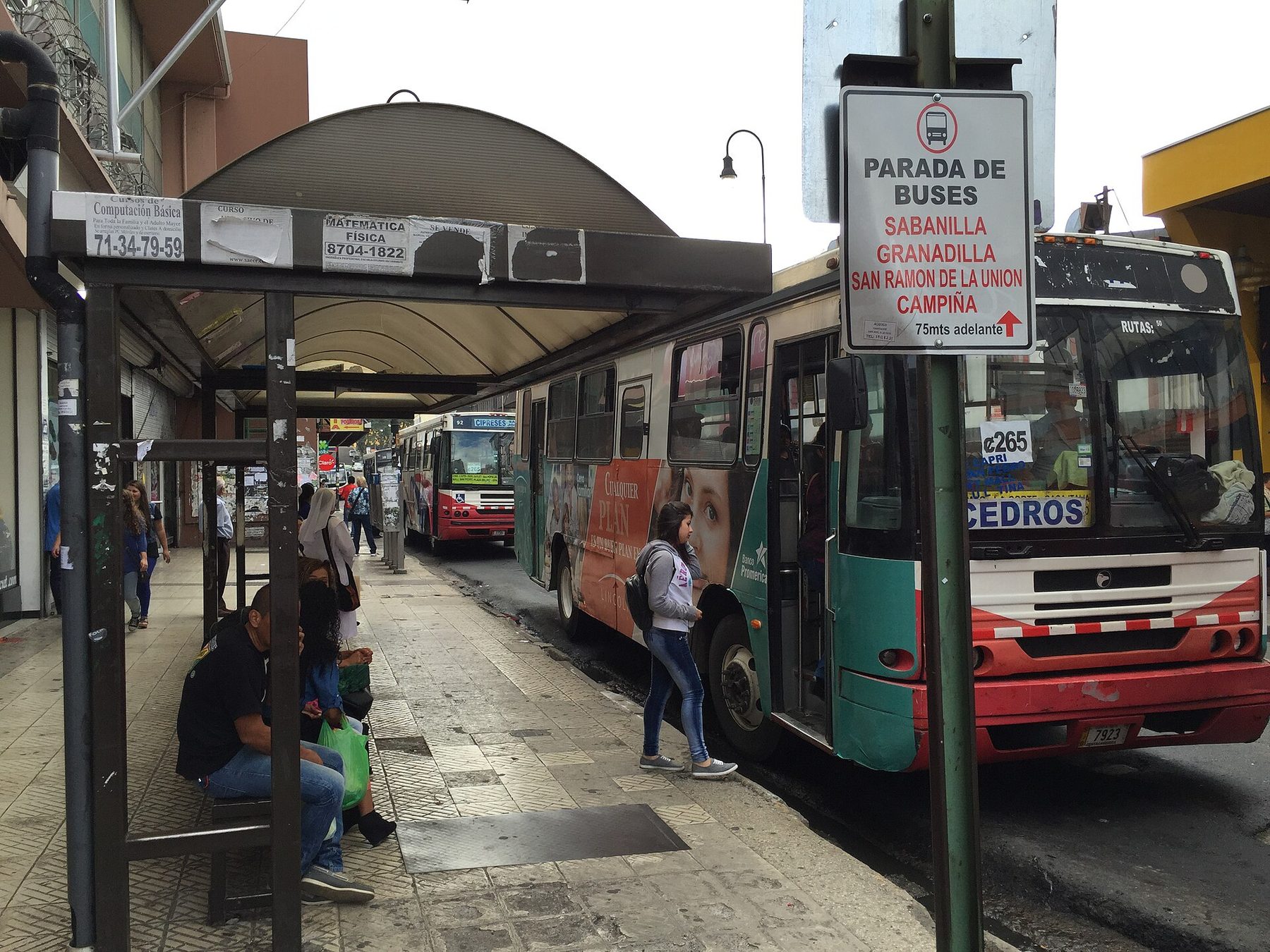 A young woman steps up into the open door of a green and red city bus at a covered pavement stop in San José, with a white sign overhead reading Parada de Buses and listing Sabanilla, Granadilla, San Ramón de la Unión and Campiña