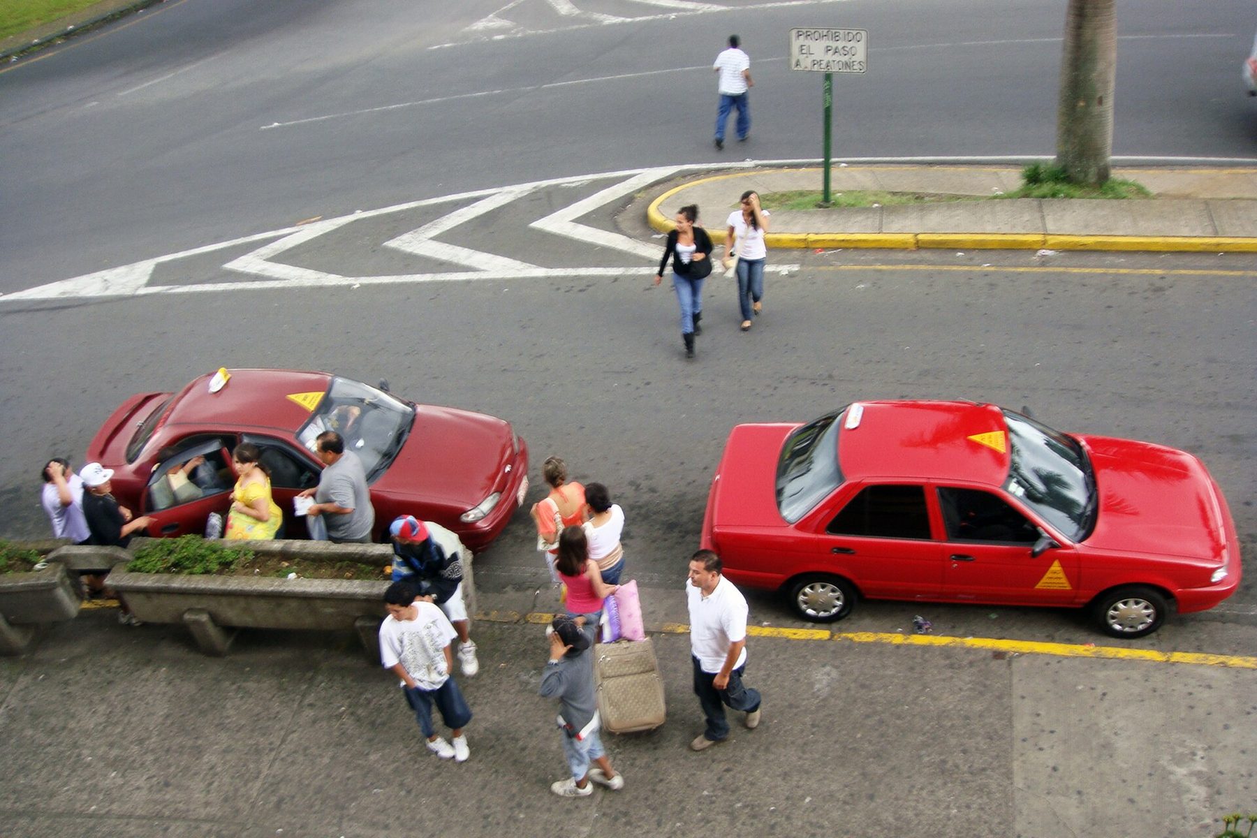 Two red taxis with yellow door triangles wait at the kerb of a wide street corner in San José while people with a wheeled suitcase and shopping bags walk past on the pavement