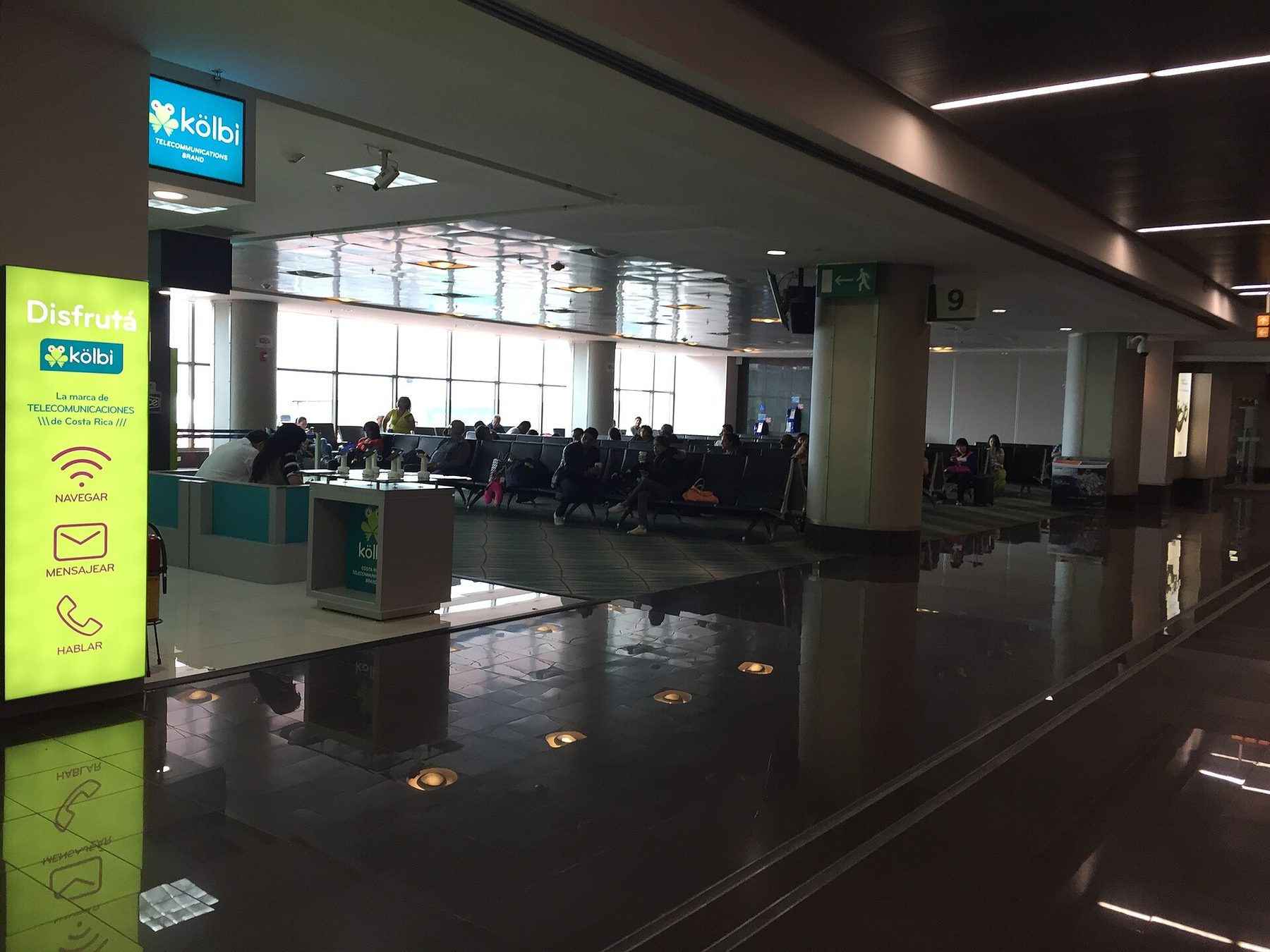 Travelers waiting in rows of seats beside gate 9 inside an airport terminal, daylight coming through tall windows and a lit advertising panel in the foreground