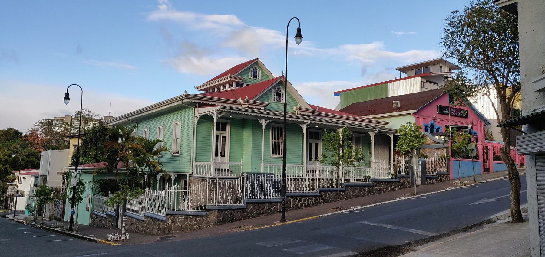 A mint-green timber house with a red roof, white veranda railings and palms in the garden, standing on a corner in Barrio Amón at dusk