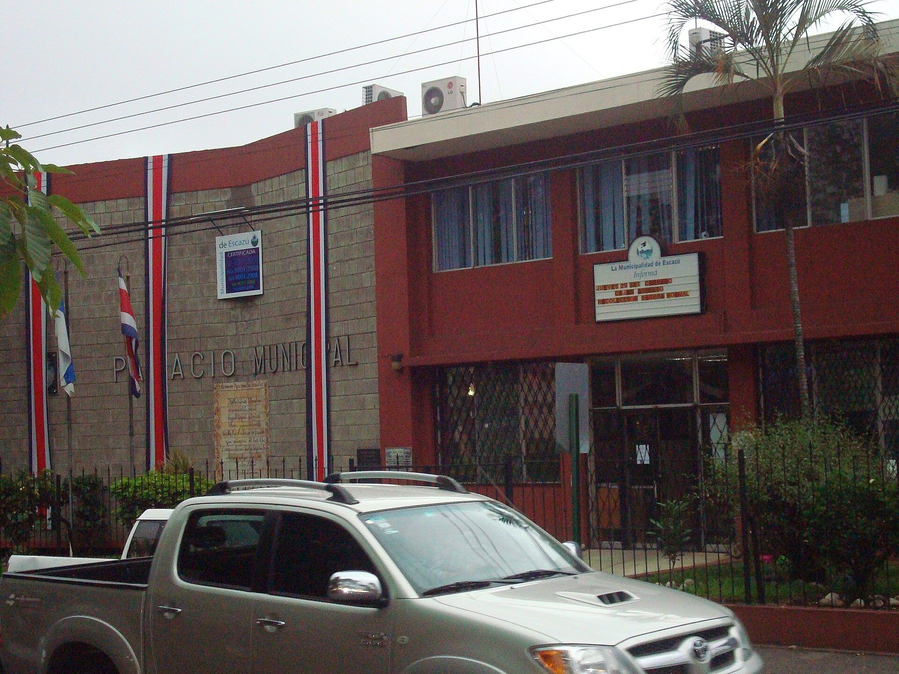 The Palacio Municipal of Escazú, a two-storey red and grey civic building with Costa Rican flags on poles and a pickup truck parked at the kerb