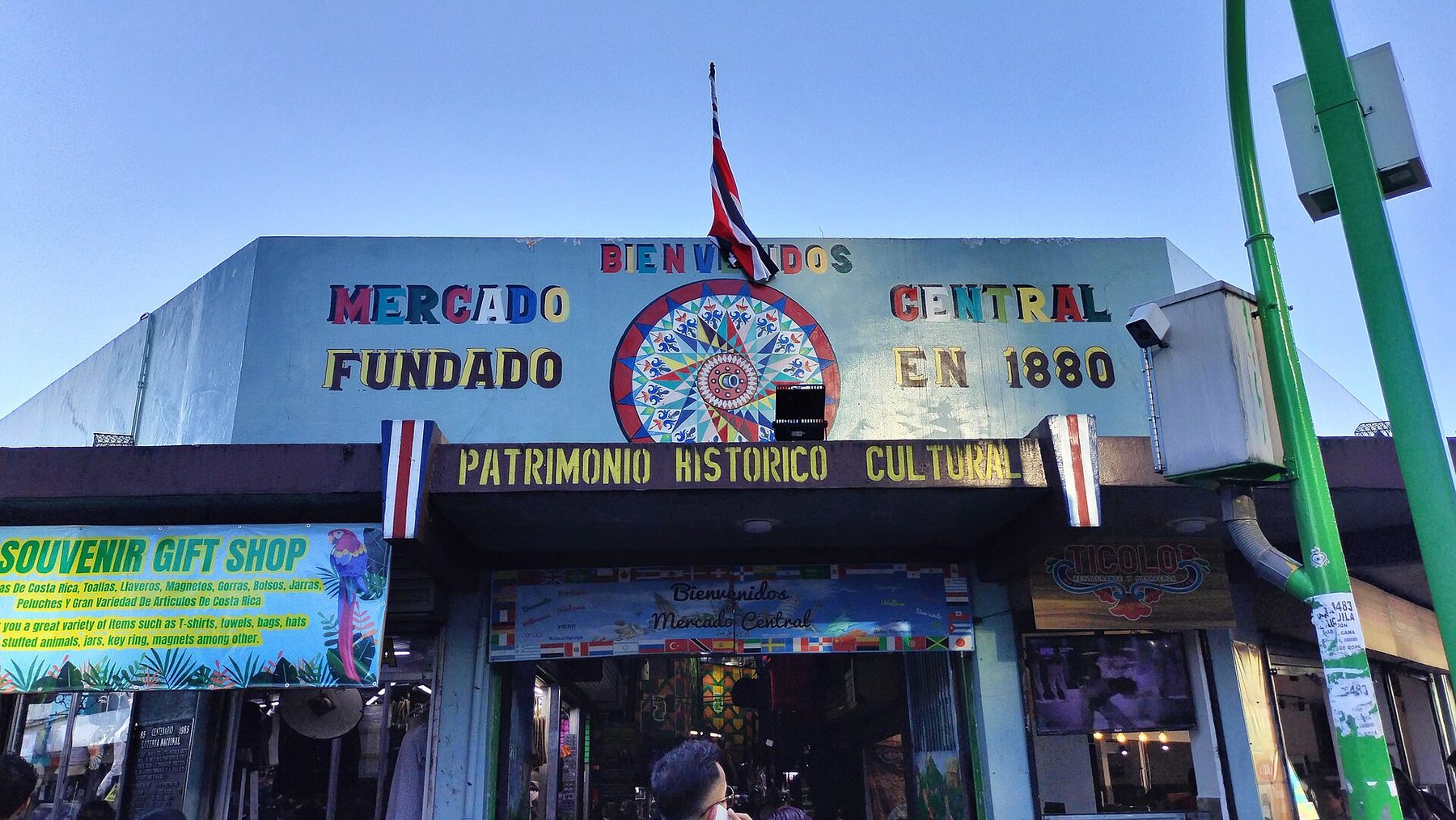 A painted blue market facade reading Mercado Central, Fundado en 1880 and Patrimonio Historico Cultural, with a Costa Rican flag above and shoppers in the doorway below