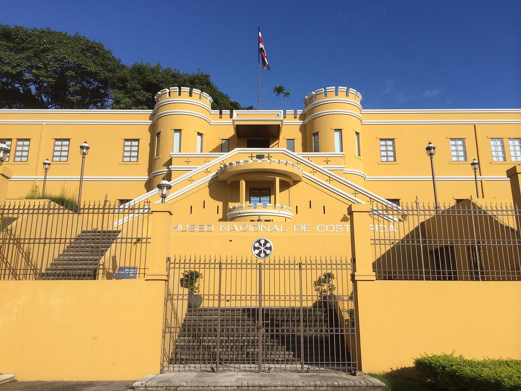 A yellow fortress-like building with two crenellated round turrets and a Costa Rican flag above, a wide double staircase, and an iron gate below lettering that reads Museo Nacional de Costa Rica