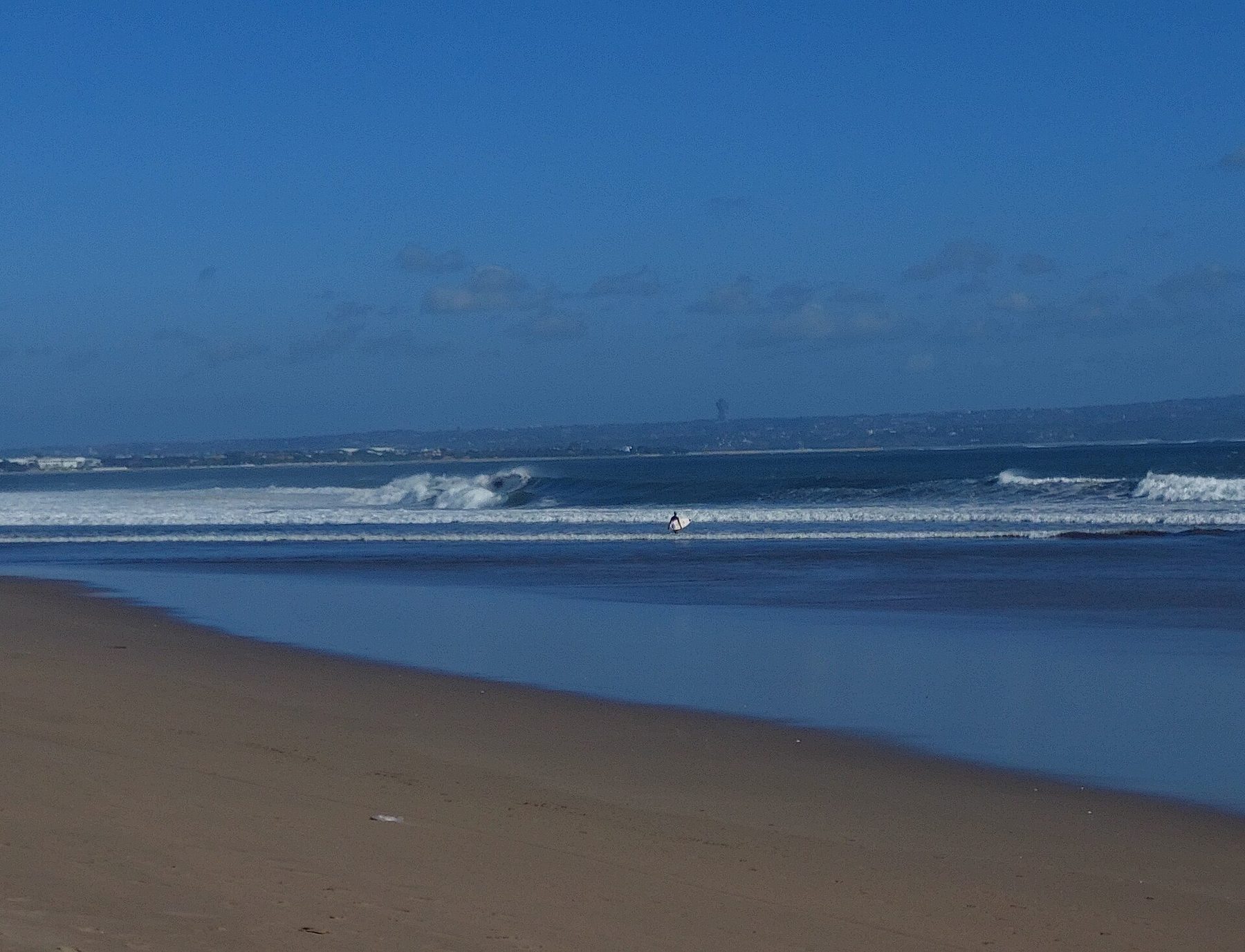 Surf running up the flat sand of Seminyak Beach with a headland visible far down the coast under a hazy sky