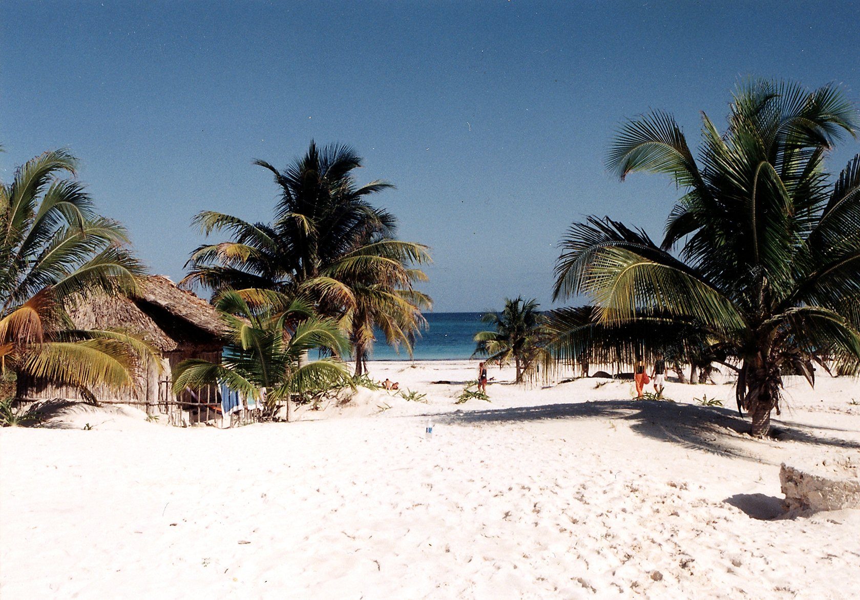 A curve of pale sand and shallow turquoise water below a low cliff, with thatched huts set back along the shore