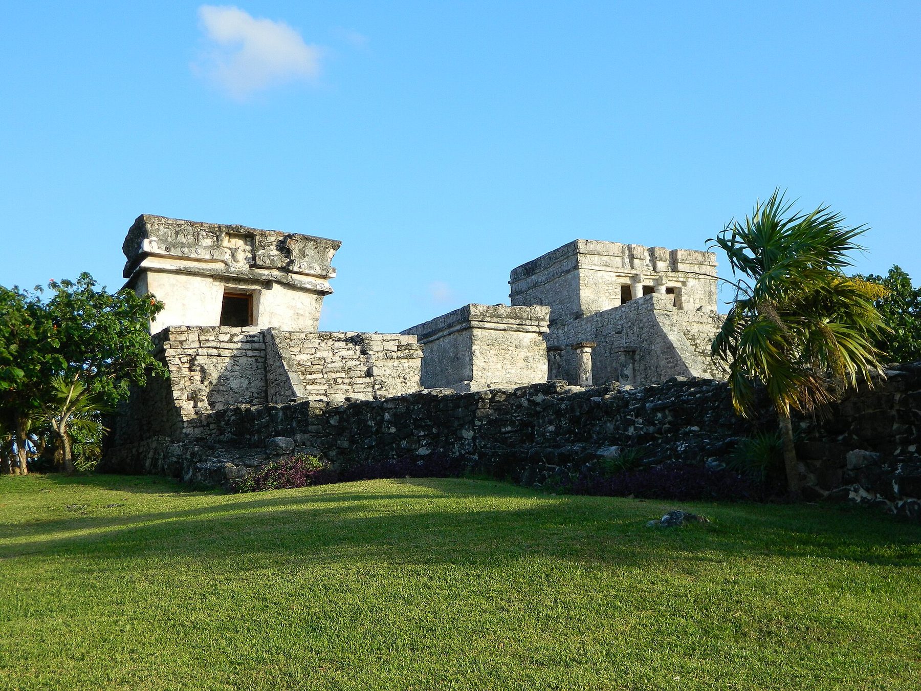 A small stone temple with a wide doorway sits on grass in front of a larger stepped pyramid under a blue sky
