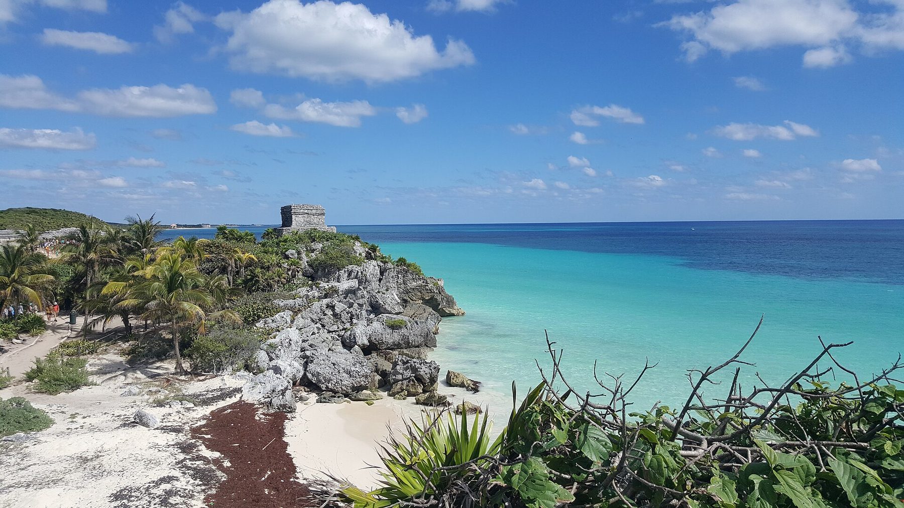 Grey stone ruins on a cliff edge above a small sand cove, with turquoise sea stretching away along the coast