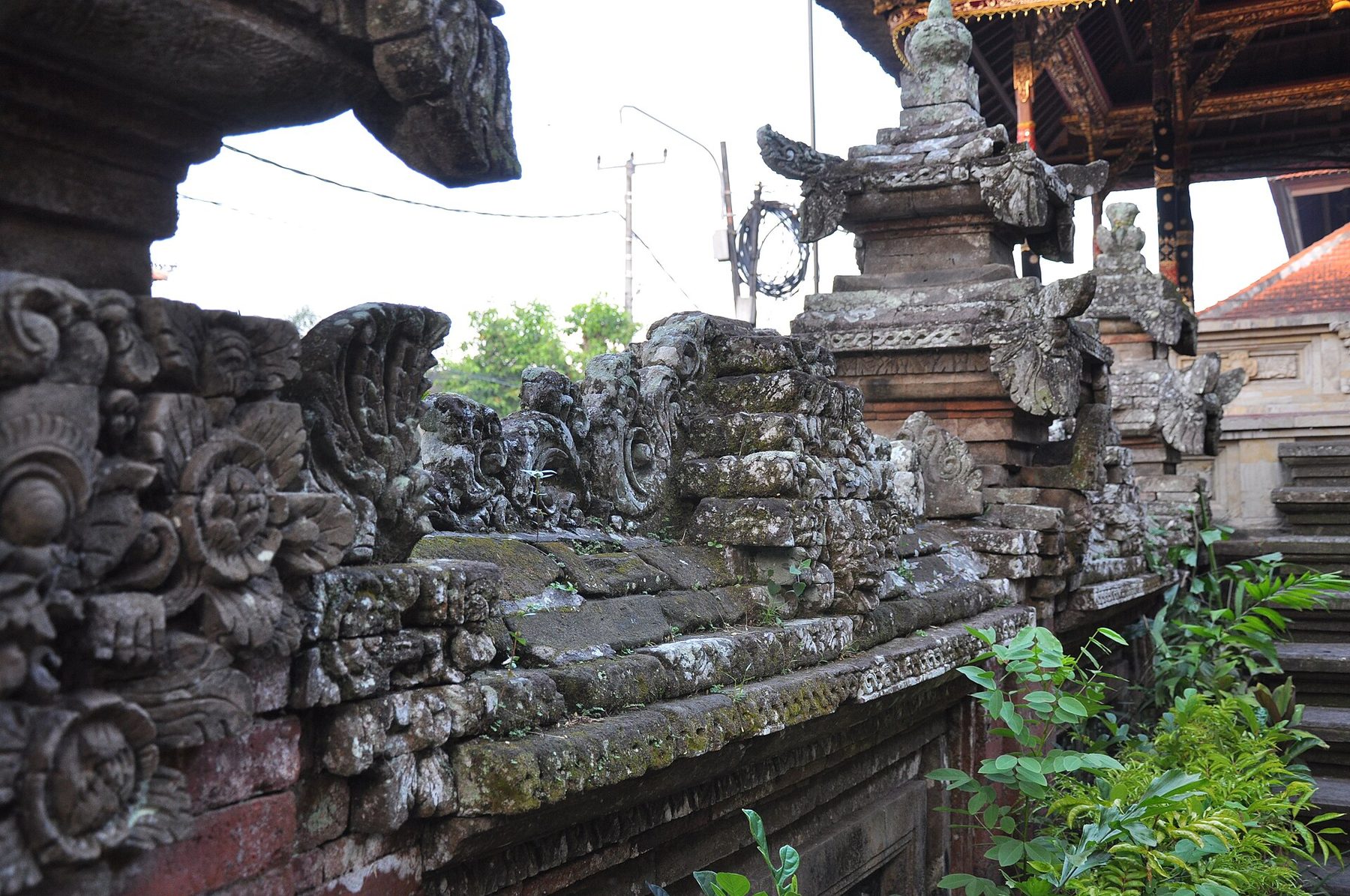 Weathered grey stone carving along a wall at Ubud Palace: floral rosettes, winged reliefs and a tiered shrine base grown over with moss and small plants, with a gilded red pavilion roof behind