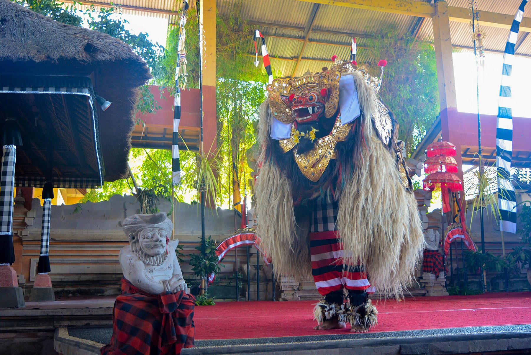 A Barong figure with a red mask, gilded crown, mirrored collar and a long pale mane stands on a red stage in an open pavilion, beside a stone guardian statue wrapped in red checked cloth