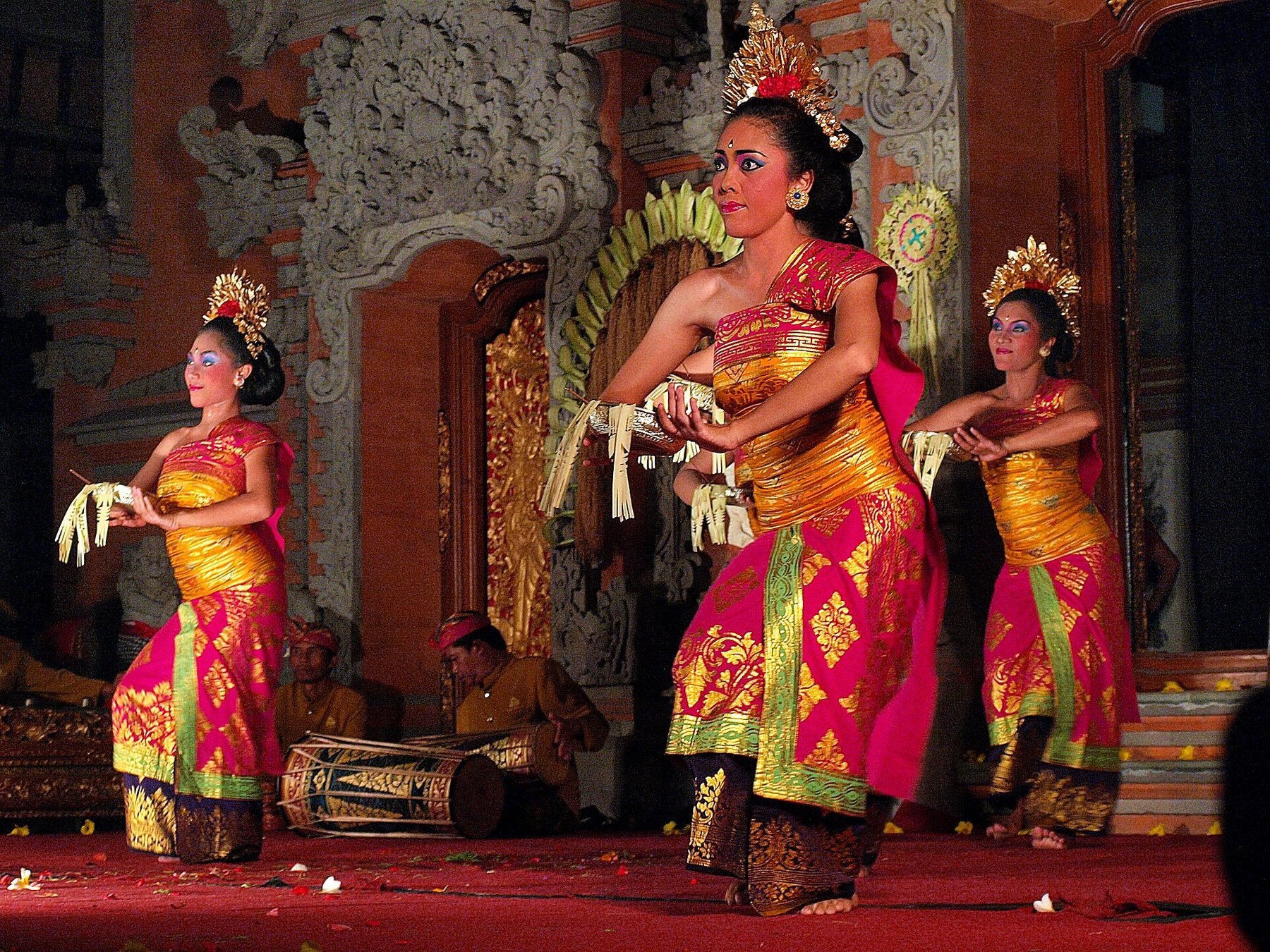 Three dancers in pink and gold sarongs and gilded headdresses hold fringed fans low across their bodies on a red stage, with two seated drummers behind them