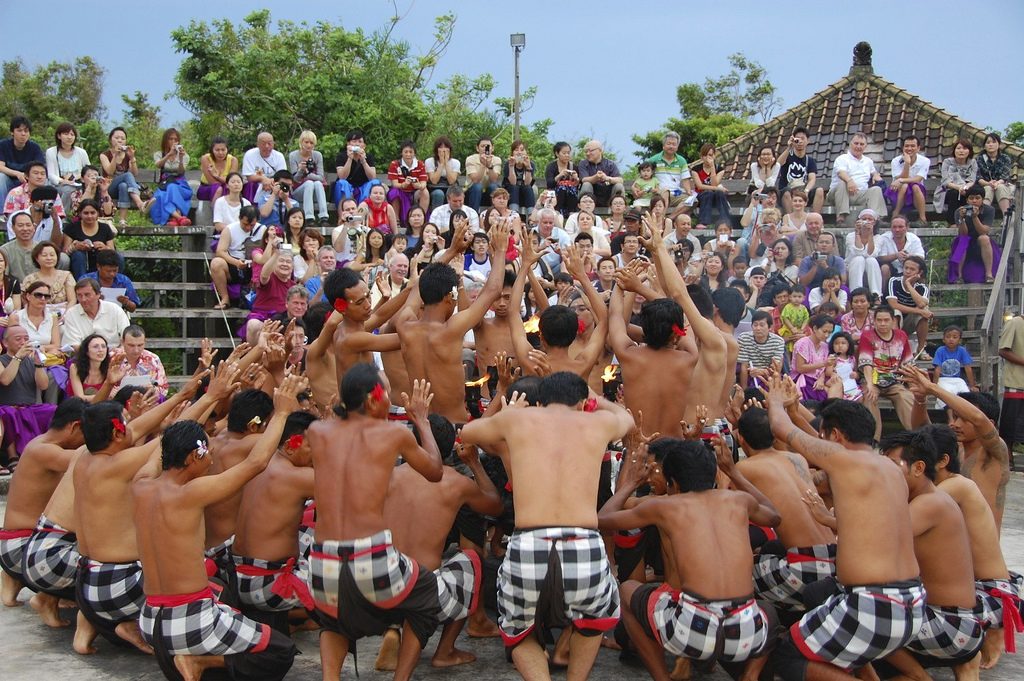 Rows of men in black and white checked cloth crouch in concentric circles with their arms raised, watched by an audience on tiered benches under a tiled temple roof