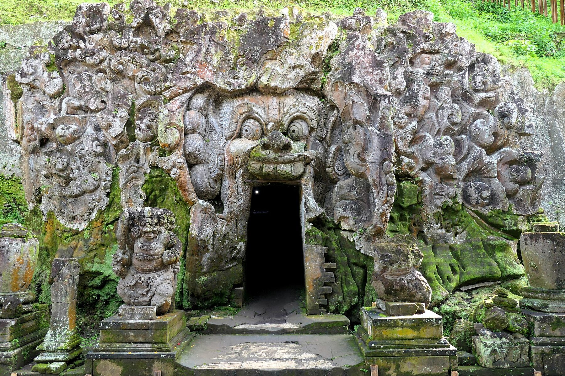 A rock face at Goa Gajah carved into a wide-eyed face with an open mouth, a dark doorway cut through the mouth, thick swirling relief carving across the whole wall and a small standing guardian statue on a plinth to the left