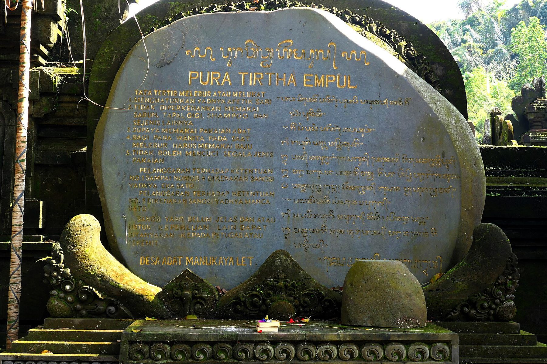 A large carved stone slab at Pura Tirta Empul with gold lettering in Indonesian and in Balinese script, listing numbered rules, signed at the foot Desa Adat Manukaya Let
