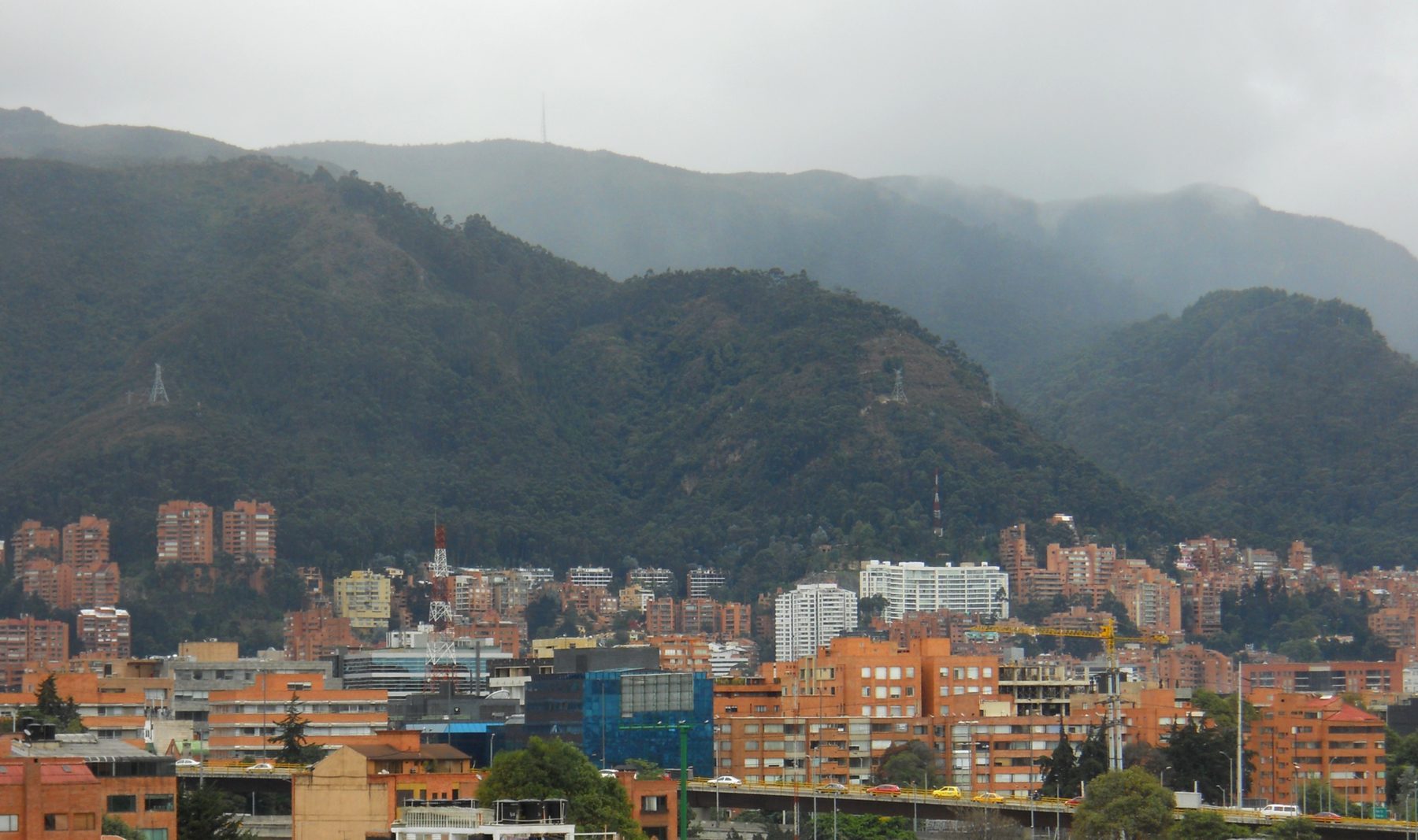 Brick apartment towers and a flyover with yellow taxis in northern Chapinero, below the forested eastern hills of Bogotá under low grey cloud