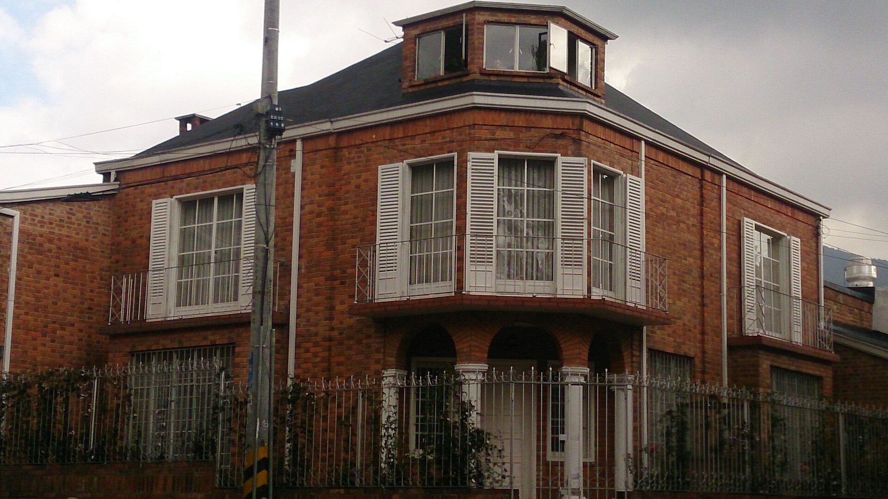 A two-storey red brick corner house in Usaquén with white shutters, wrought-iron balconies and railings, ivy along the garden wall and a green hill in the distance
