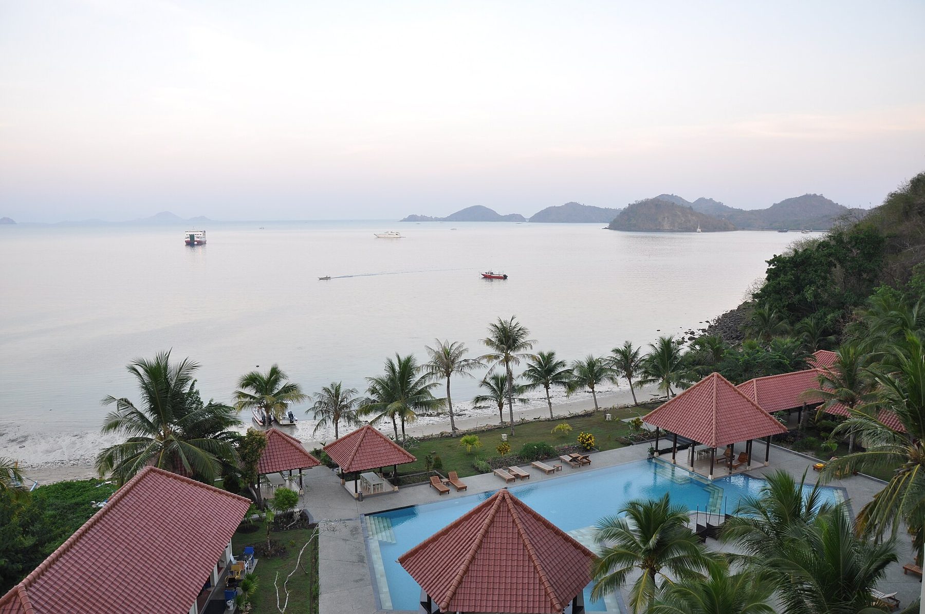 A dusk view from high ground over a bay at Labuan Bajo, looking down on red-tiled pavilion roofs, a swimming pool, palms and a pale beach, with a ferry and low islands on the water beyond