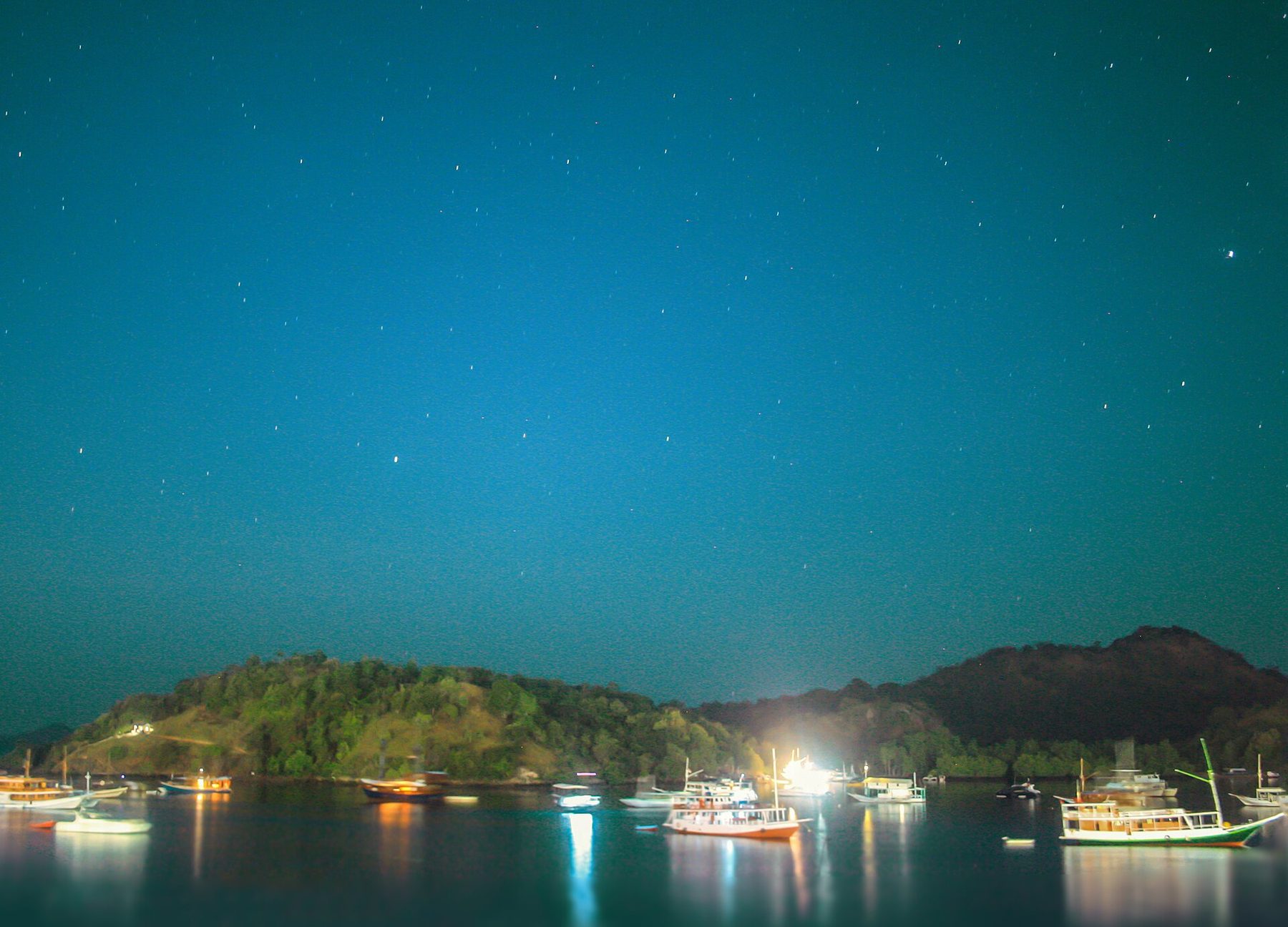 Fishing and tour boats lying at anchor off Labuan Bajo at night under a star-filled sky, deck lights reflecting in flat water, with dark wooded headlands behind