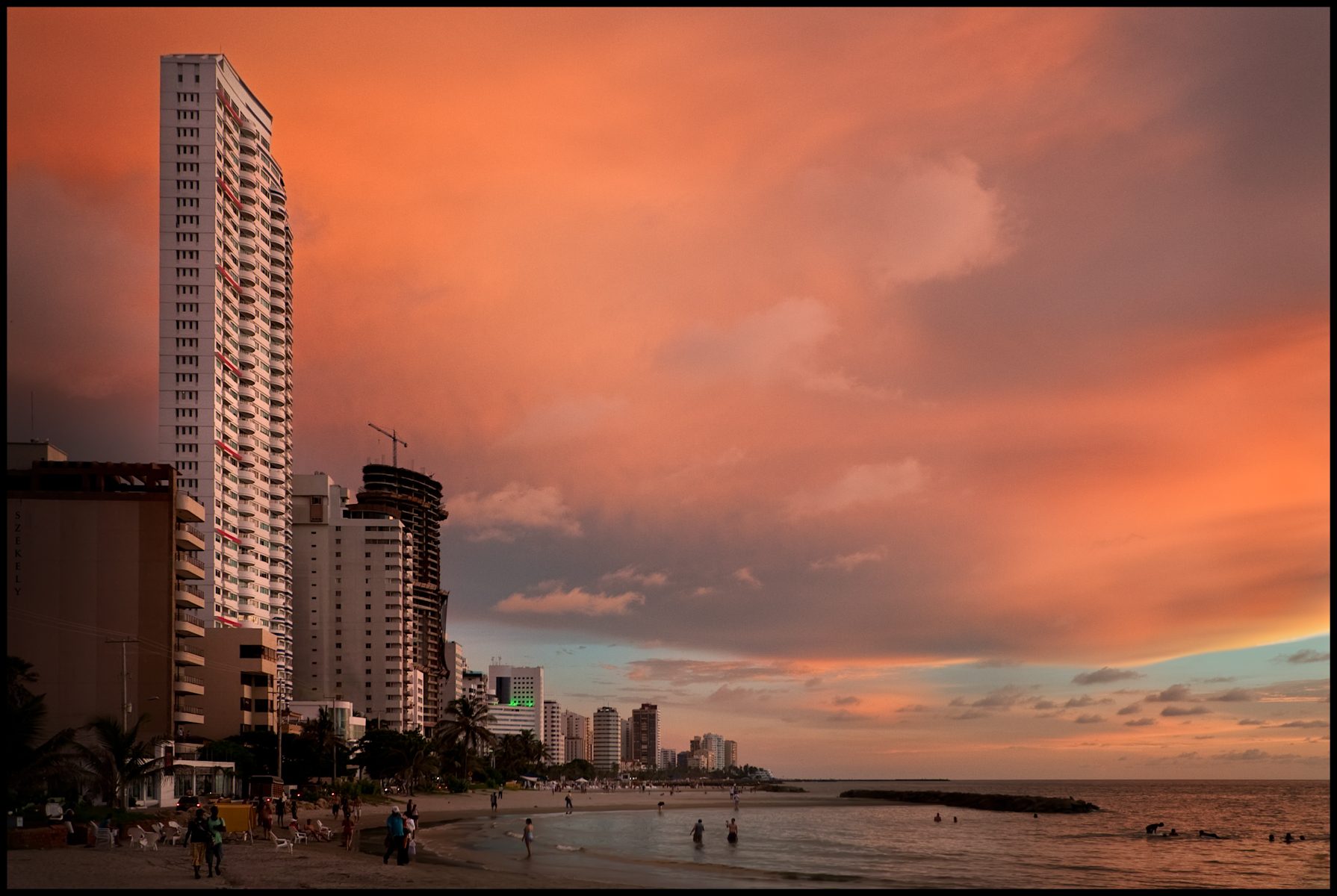 A wide grey-sand beach at Bocagrande with striped umbrellas and sun loungers in rows, the Caribbean on one side and a line of white apartment towers on the other