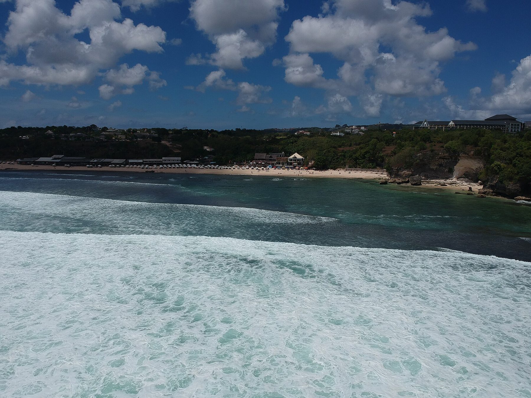 A wide sandy beach at Balangan backed by low cliffs and palm-thatched warungs, with surf rolling in from the left