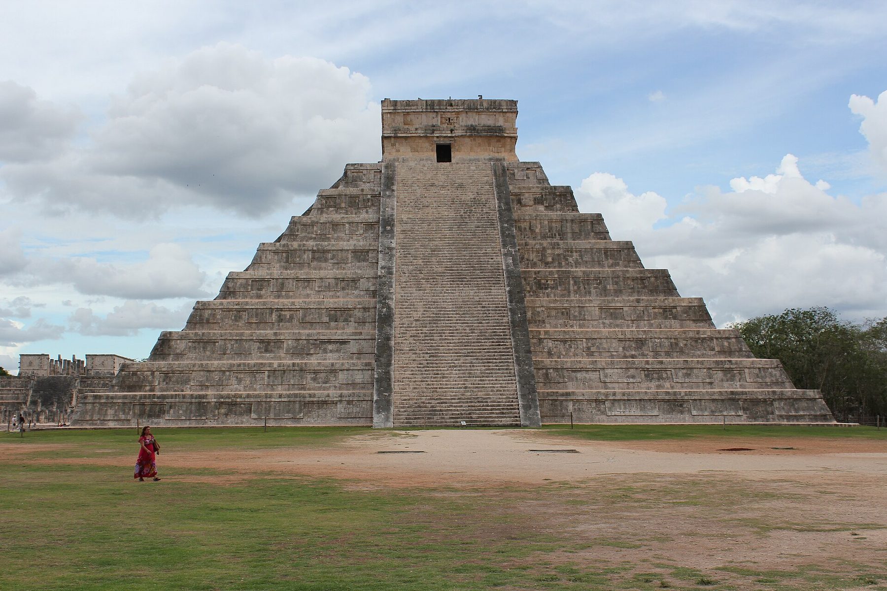 A stepped stone pyramid with a temple on its flat top and a wide stairway down the near face, standing on mown grass under a pale sky