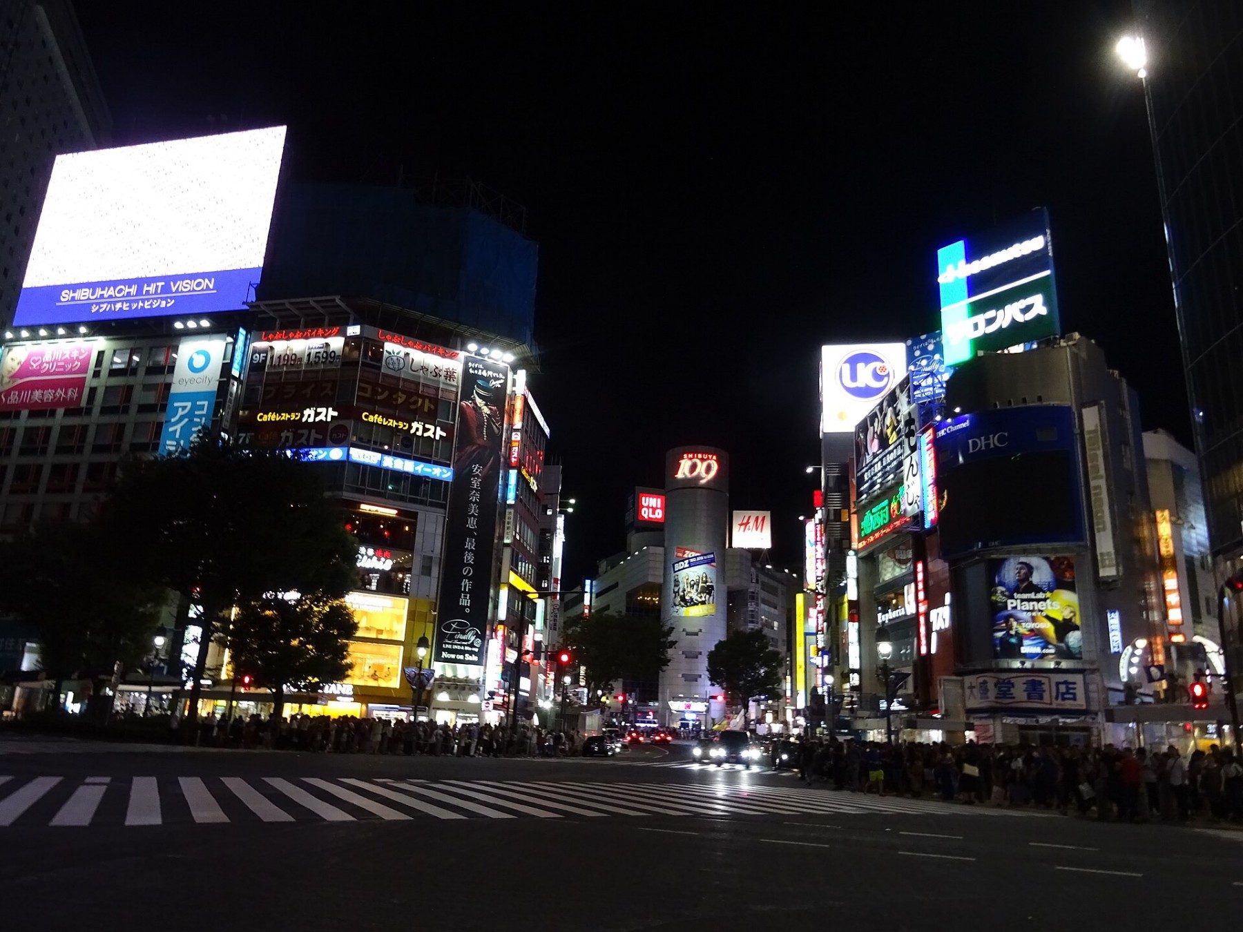 Nighttime crowds crossing broad striped crosswalks beneath illuminated buildings in Shibuya, Tokyo